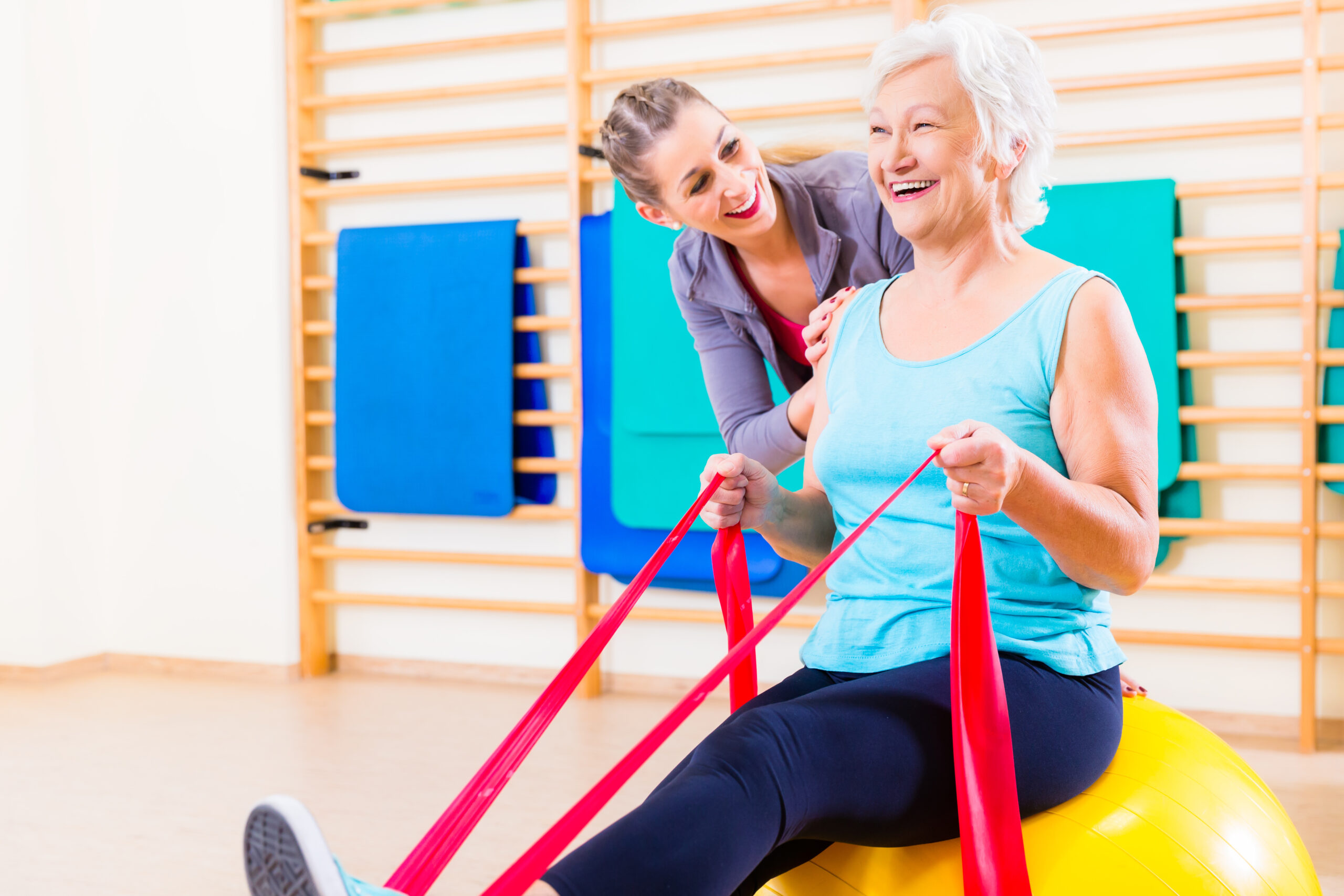 Senior woman with stretch band in fitness gym being coached by personal trainer